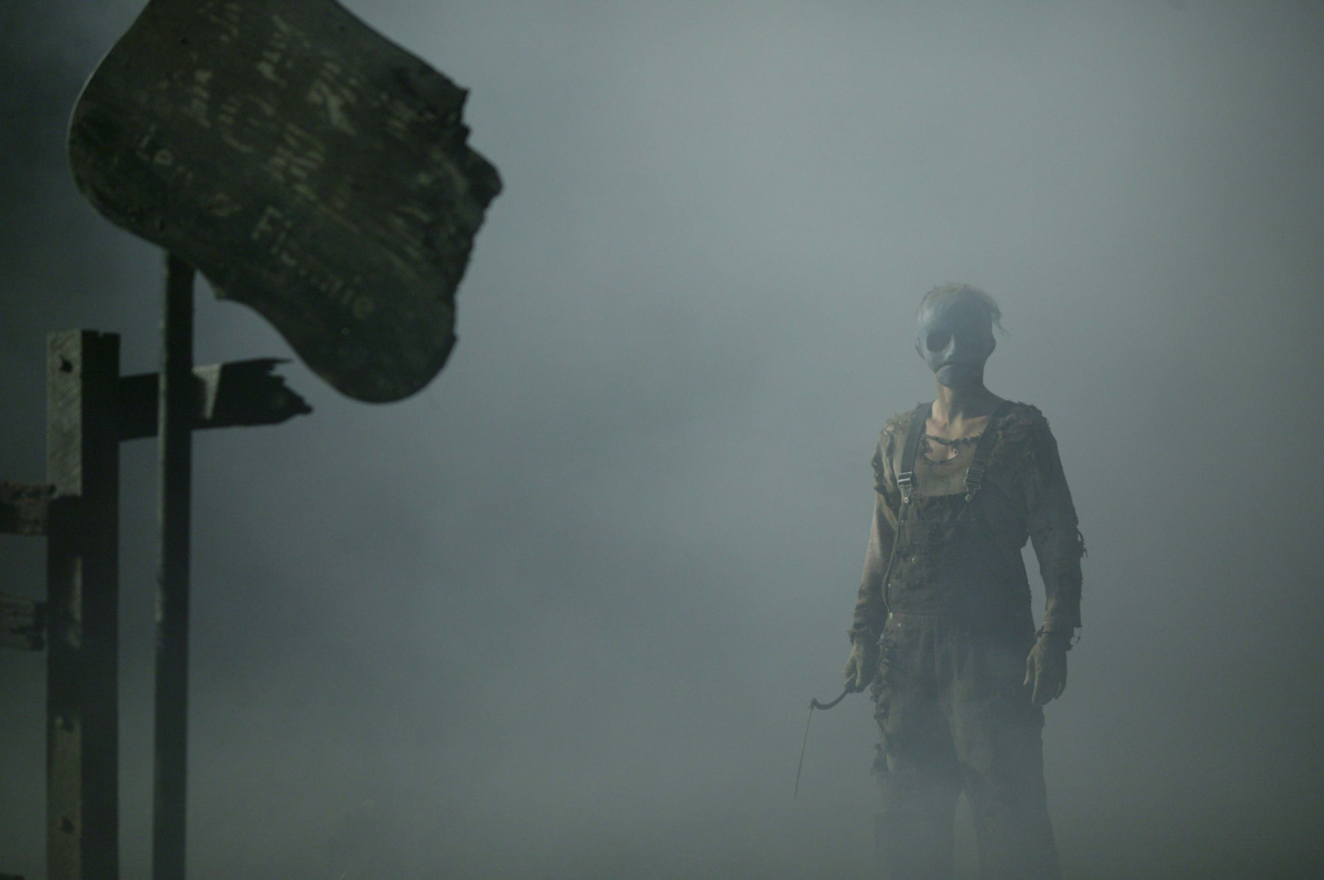 Leslie Vernon, masked and in worn work clothes, standing in heavy fog beside a weathered wooden sign